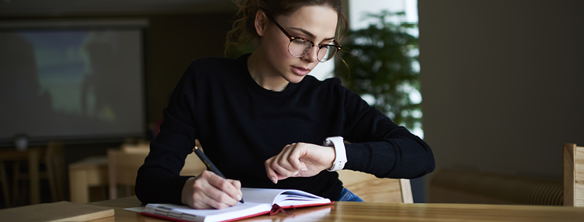 Symbolbild Zeitmanagment: Frau mit Brille sitzt an einem Tisch, schreibt etwas und schaut auf Armbanduhr